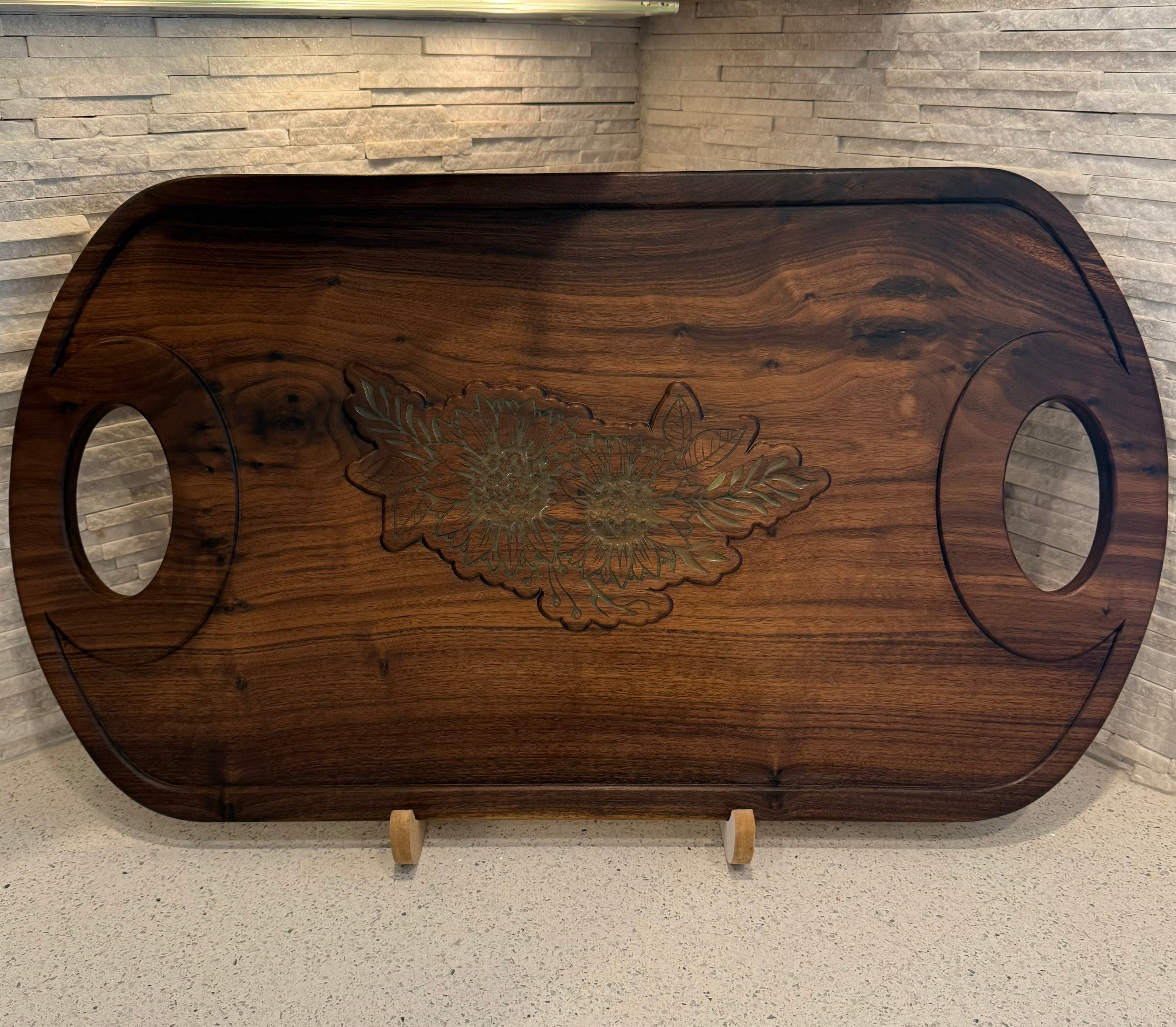 Wooden cutting board with leaf design on a stand against a stone wall background